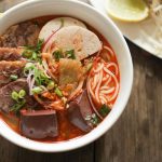 Tô bún bò Huế đầy ắp thịt bò, chả, huyết kèm đĩa rau sống và chanh tươi trên bàn gỗ